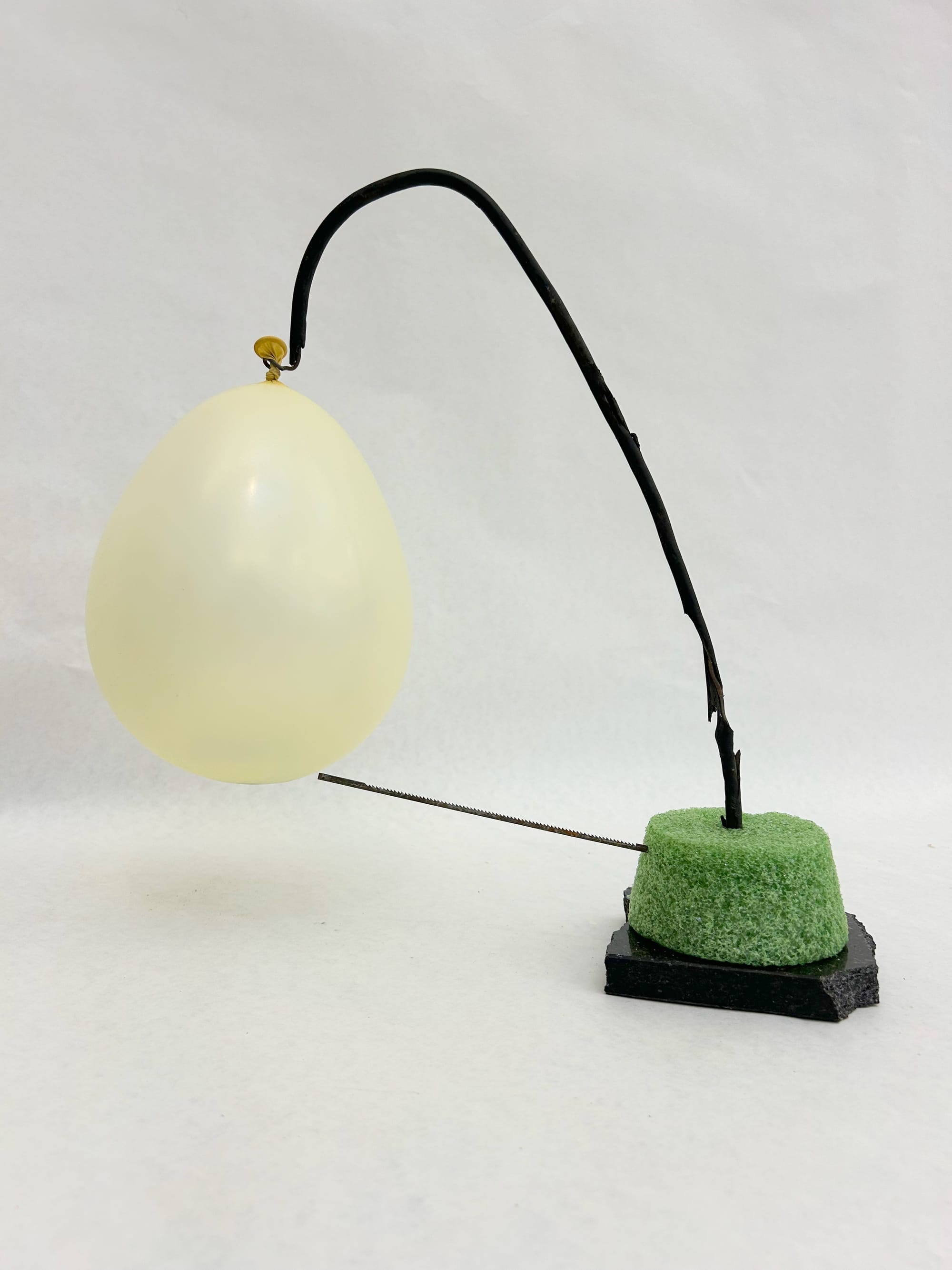 a small sculpture with a black marble base with green styrofoam on top, which has a worn piece of rubber coated wire coming out of it, and stretching up and away. From the wire there is a balloon filled with marbles hanging right over a sawblade