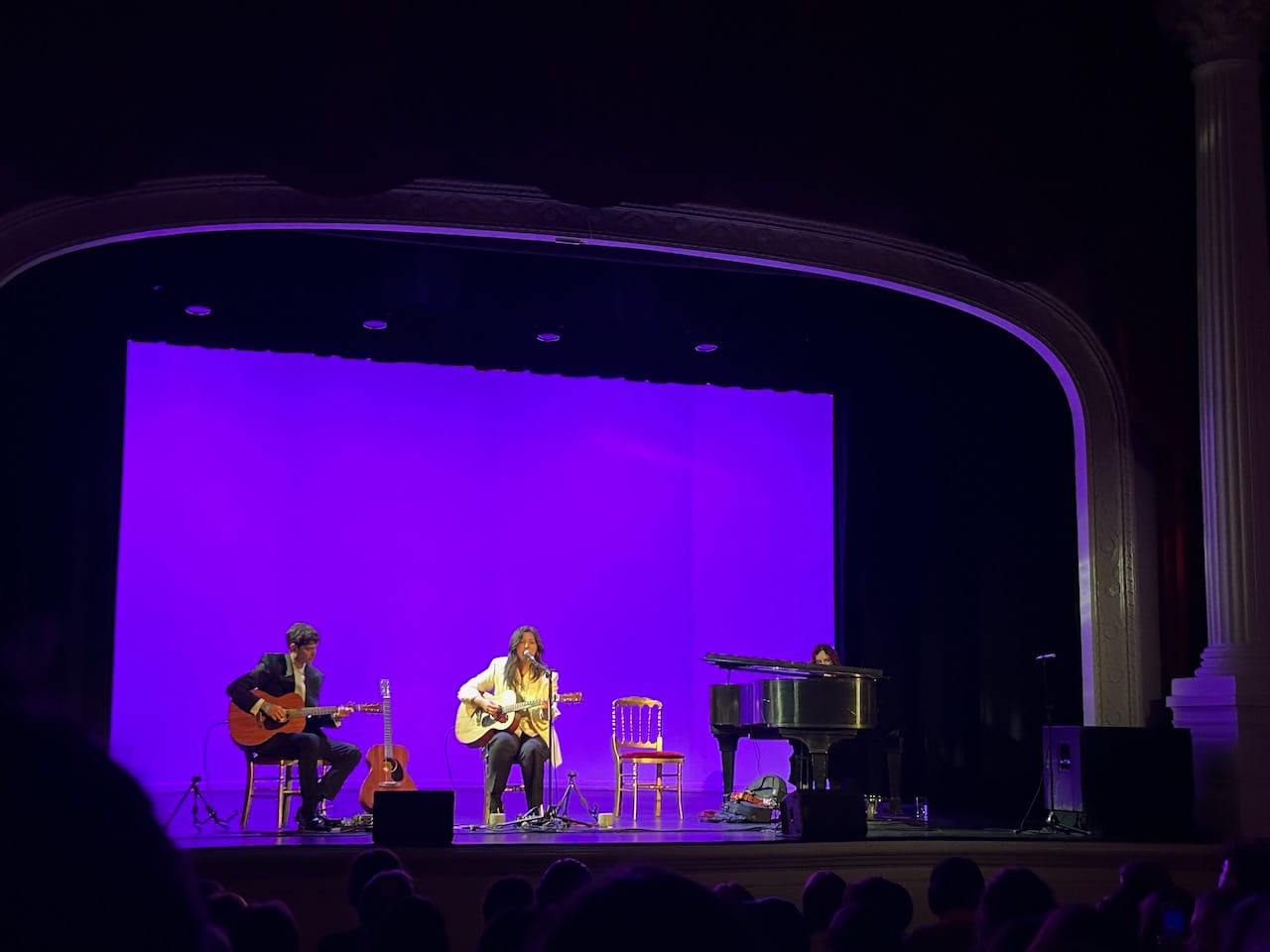 A stage backlit with purple light. There are three musicians, a man on guitar, a woman on guitar, and a woman on piano. Lucy Dacus is the middle person, wearing a white blazer.